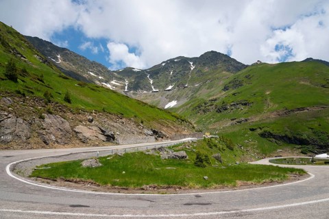 Fresh green hills and winding mountain road, partly covered by snow under a partly cloudy sky,