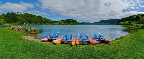 Tranquil Furnas Lake with pedal boats on the shore, surrounded by green nature and a cloudy sky,