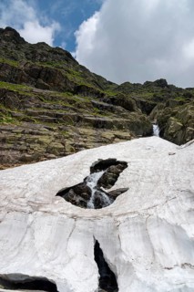 Snow-covered mountain landscape with rocks and a stream under a cloudy sky, small Capra waterfall,