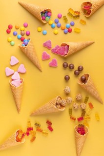 Colourful sweets, gummy bears and chocolate lentils fall out of ice cream cones on a yellow