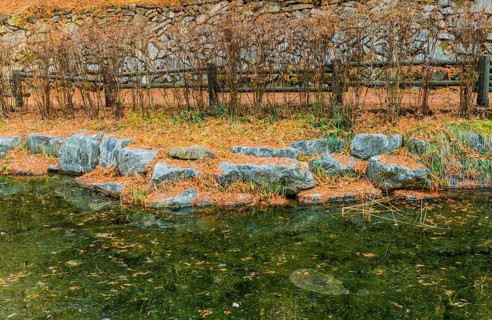 Boulders on shoreline of small frozen pond with wooden railing fence and stone wall in background