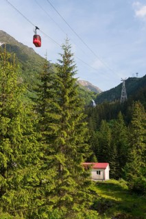 Small cable car gondola crosses a picturesque mountain landscape with fir trees and a small hut and