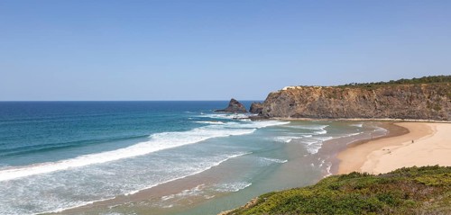 Atlantic Ocean waves breaking on rocky headland and bay with wide sandy beach, surfer and a few