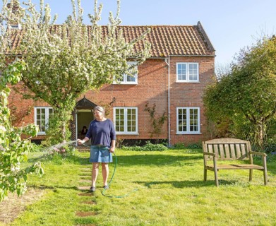 Woman watering garden with hosepipe in front of red brick historic detached house, Shottisham,