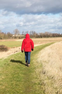 Woman wearing red winter jacket walking on raised coastal flood defence dyke embankment, Hollesley