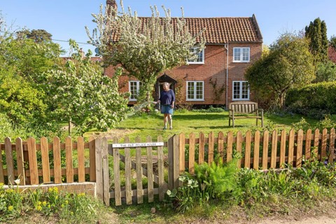 Woman watering garden with hosepipe in front of red brick historic detached house, Shottisham,