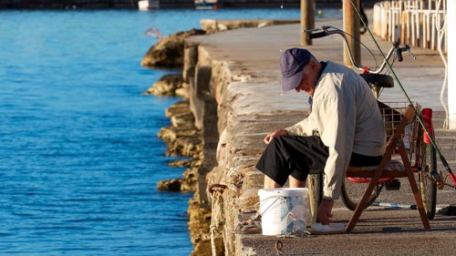 A man sitting on a folding chair fishing on the waterfront in the morning light, Gythio, Mani,