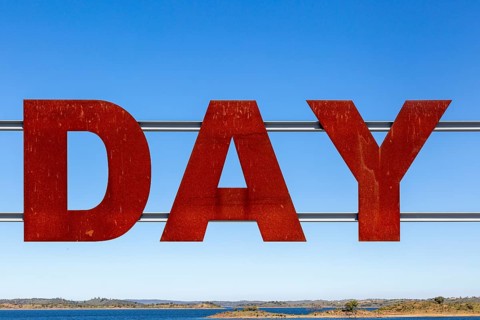 Large red rusty capital letters spell the word DAY against blue sky background, Alqueva dam, Moura,