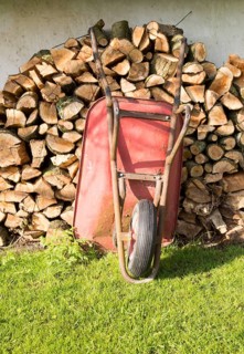 Old red metal wheelbarrow leaning against pile wood logs in garden, Cherhill, Wiltshire, England,