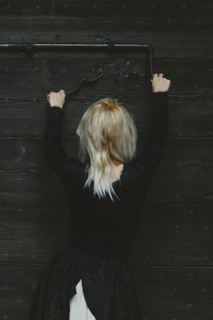 Back view of a blonde thin white woman dressed in black reaching up to a rusty antique metal lock