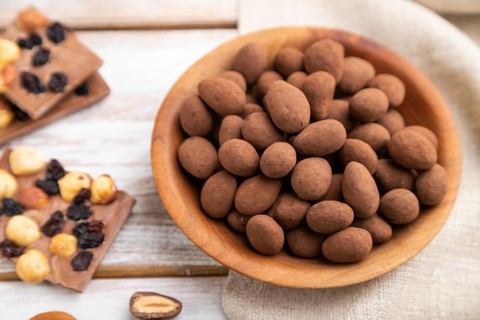 Almond in chocolate dragees in wooden plate and a cup of coffee on white wooden background and