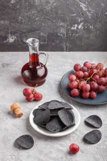 Black potato chips with charcoal, balsamic vinegar in glass, red grapes on a blue ceramic plate on