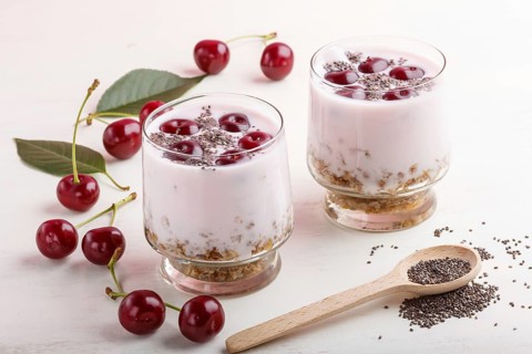 Yoghurt with cherries, chia seeds and granola in glass with wooden spoon on white wooden background