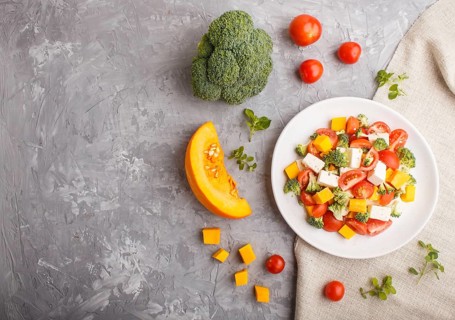 Vegetarian salad with broccoli, tomatoes, feta cheese, and pumpkin on white ceramic plate on a gray