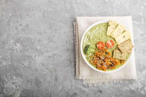 Green broccoli cream soup with crackers and cheese in white bowl on a gray concrete background and
