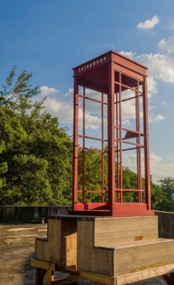 Red wooden telephone booth minus phone and without glass on roof of building on sunny day with blue