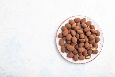 Almond in chocolate dragees on ceramic plate on white concrete background. Top view, copy space,