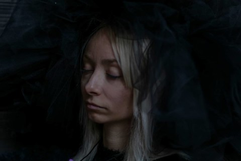 A young caucasian blonde woman's face is partially obscured by a dark veil, her eyes closed in a