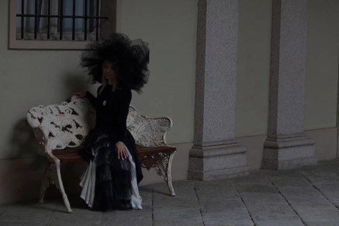 Thoughtful woman in black gothic attire sitting on a bench by a historical building, wear lace veil