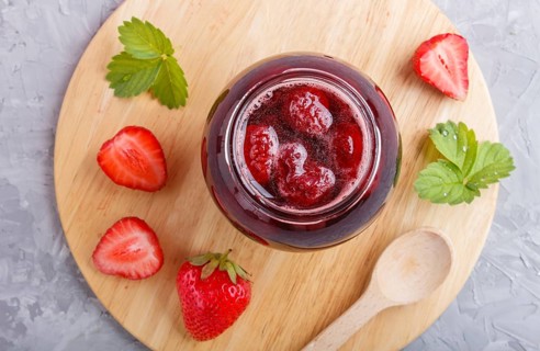 Strawberry jam in a glass jar with berries and leaves on gray concrete background. Homemade, top