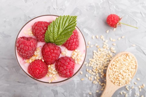 Yoghurt with raspberry and sesame in a glass and wooden spoon on gray concrete background. top