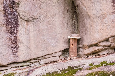 Log held in place with stones and carved to have one eye and mouth on rocky ledge of cliff face in