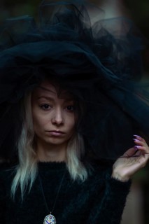 Mysterious white young blonde woman in a dark setting wear a large black tulle hat and veil and a