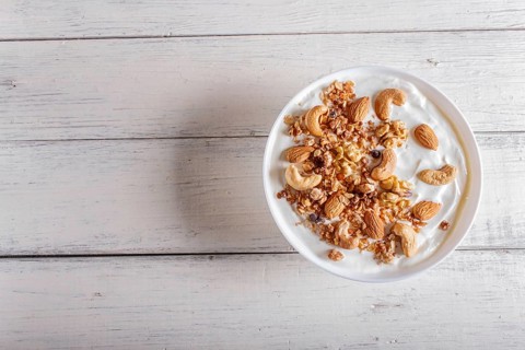 White plate with greek yogurt, granola, almond, cashew, walnuts on white wooden background. top