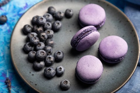 Purple macarons or macaroons cakes with blueberries on ceramic plate on a blue concrete background