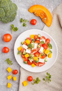 Vegetarian salad with broccoli, tomatoes, feta cheese, and pumpkin on white ceramic plate on a gray