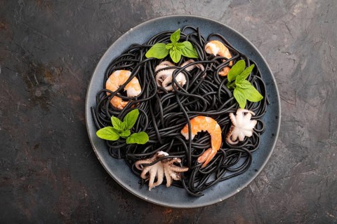 Black cuttlefish ink pasta with shrimps or prawns and small octopuses on black concrete background.