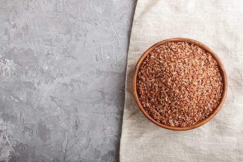 Wooden bowl with unpolished brown rice on a gray concrete background and linen textile. Top view,