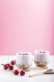 Yoghurt with cherries, chia seeds and granola in glass with wooden spoon on pink and white
