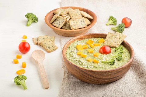 Green broccoli cream soup with crackers and cheese in wooden bowl on a white wooden background and