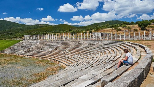 Person sitting in ancient stadium under clear blue sky with mountains in the background,