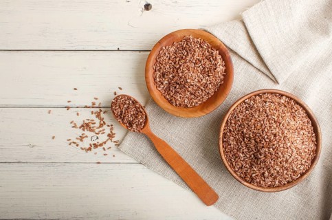 Two wooden bowls with unpolished brown rice and wooden spoon on a white wooden background and linen