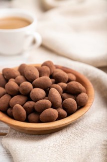 Almond in chocolate dragees in wooden plate and a cup of coffee on white wooden background and