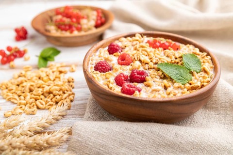 Wheat flakes porridge with milk, raspberry and currant in wooden bowl on white wooden background