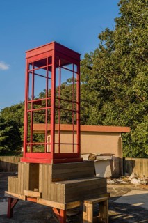 Red wooden telephone booth minus phone and without glass on roof of building on sunny day with blue