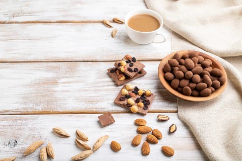 Almond in chocolate dragees in wooden plate and a cup of coffee on white wooden background and