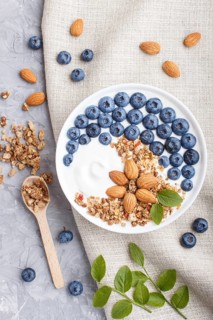 Yoghurt with blueberry, granola and almond in white plate with wooden spoon on gray concrete