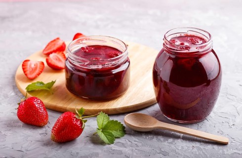 Strawberry jam in a glass jar with berries and leaves on gray concrete background. Homemade, close