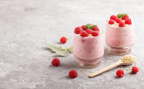 Yoghurt with raspberry and sesame in a glass and wooden spoon on gray concrete background. side