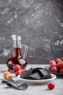 Black potato chips with charcoal, balsamic vinegar in glass, red grapes on a blue ceramic plate on