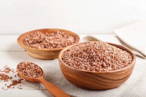 Two wooden bowls with unpolished brown rice and wooden spoon on a white wooden background and linen