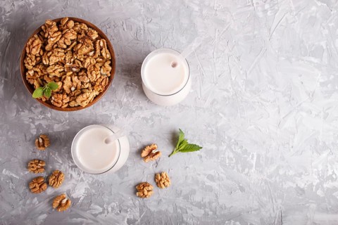 Organic non dairy walnut milk in glass and wooden plate with walnuts on a gray concrete background.