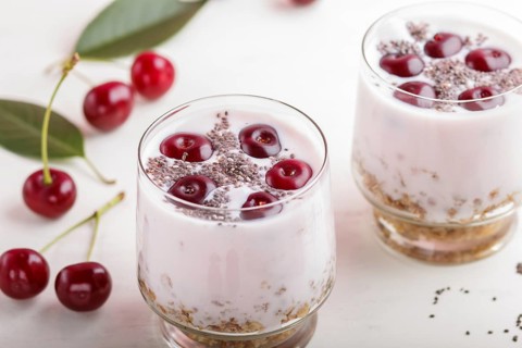 Yoghurt with cherries, chia seeds and granola in glass with wooden spoon on white wooden background