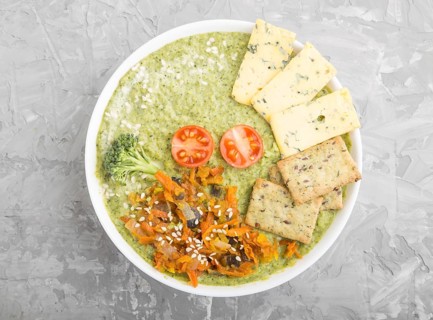 Green broccoli cream soup with crackers and cheese in white bowl on a gray concrete background. top