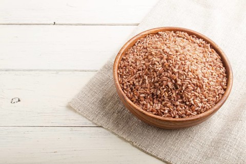 Wooden bowl with unpolished brown rice on a white wooden background and linen textile. Side view,