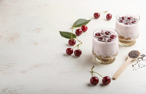 Yoghurt with cherries, chia seeds and granola in glass with wooden spoon on white wooden background